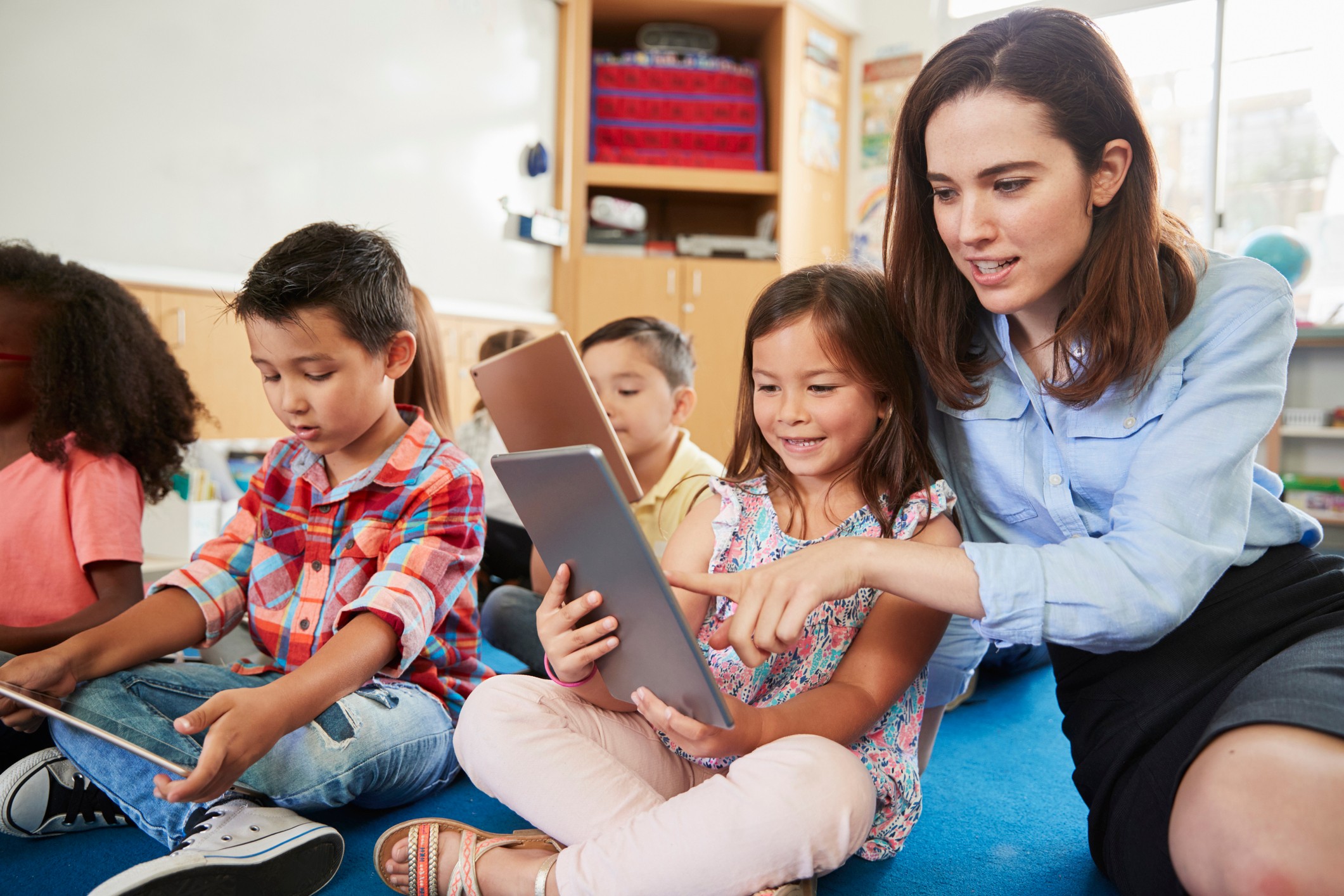 Teacher works with group of children sitting on the floor and playing Prodigy on iPads.