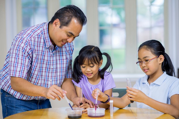 A father helps his two young daughters with an at-home science experiment.