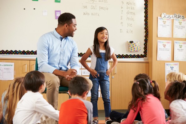 A student stands at the front of the classroom with her teacher, talking to her classmates, who are seated.