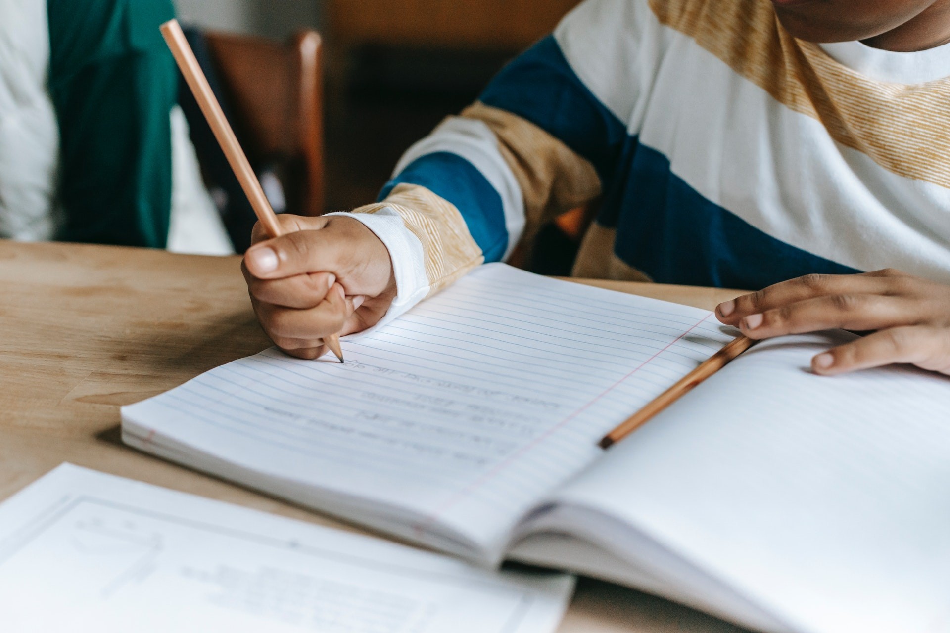 Young child sits at a desk with a notebook and pencil, writing in the notebook.