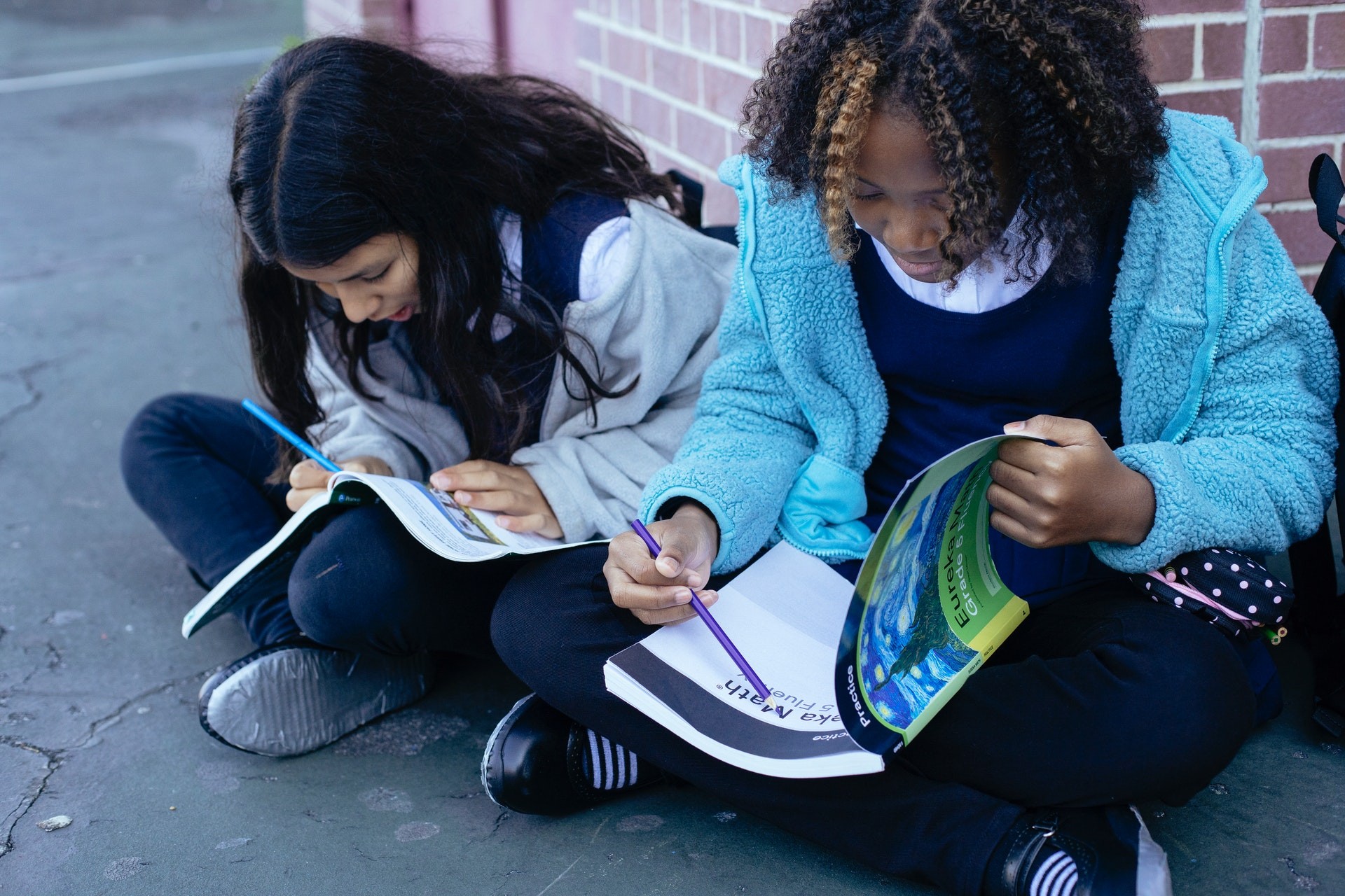 Two students sit outside against a brick wall, working in notebooks.