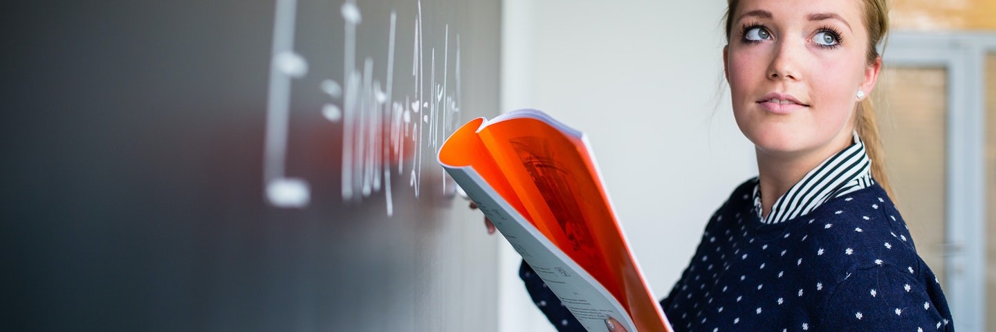 A teacher stands at the blackboard and writes a math word problem while holding an orange workbook.