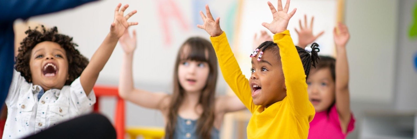 Young students sit on a mat in the classroom and play classroom games with their teacher.
