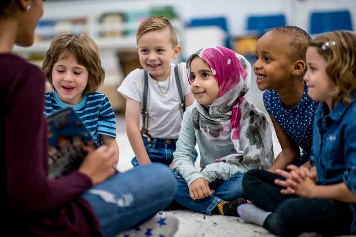 Five students sit cross legged on the ground in front of a teacher holding a book.