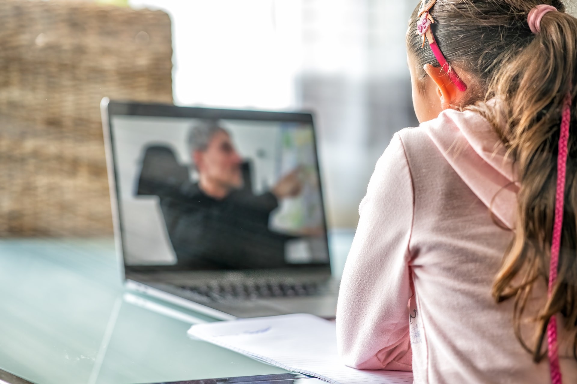 Young student sits at a table and watches her teacher on the screen during virtual learning.
