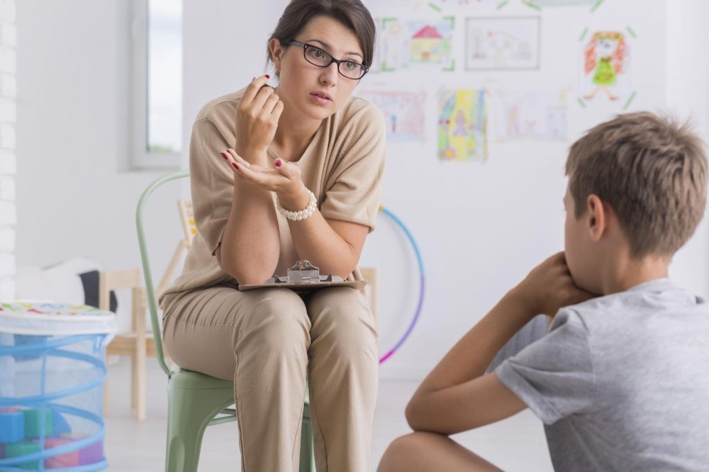 A female teacher sitting on a chair is carefully listening to a student
