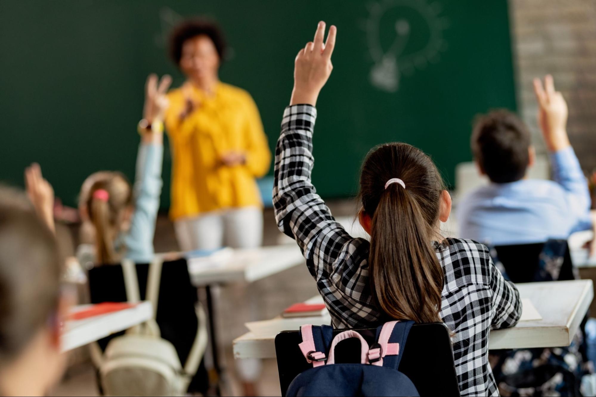 Teacher stands at the front of the classroom and points as students as they raise their hands after returning to in-person learning.