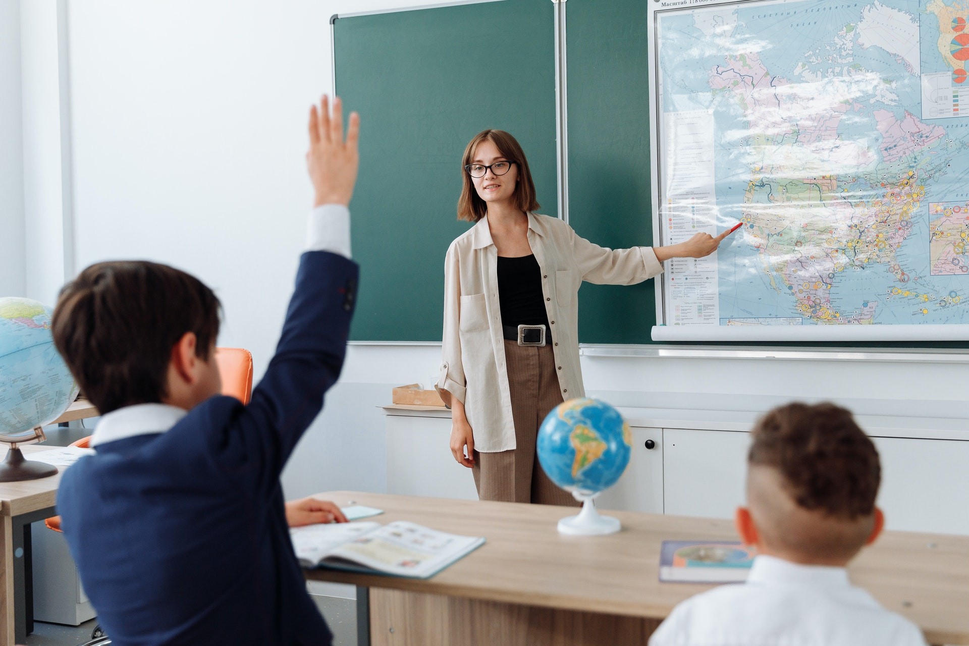 A teacher stands at the front of a classroom and points at a map in front of the chalkboard.