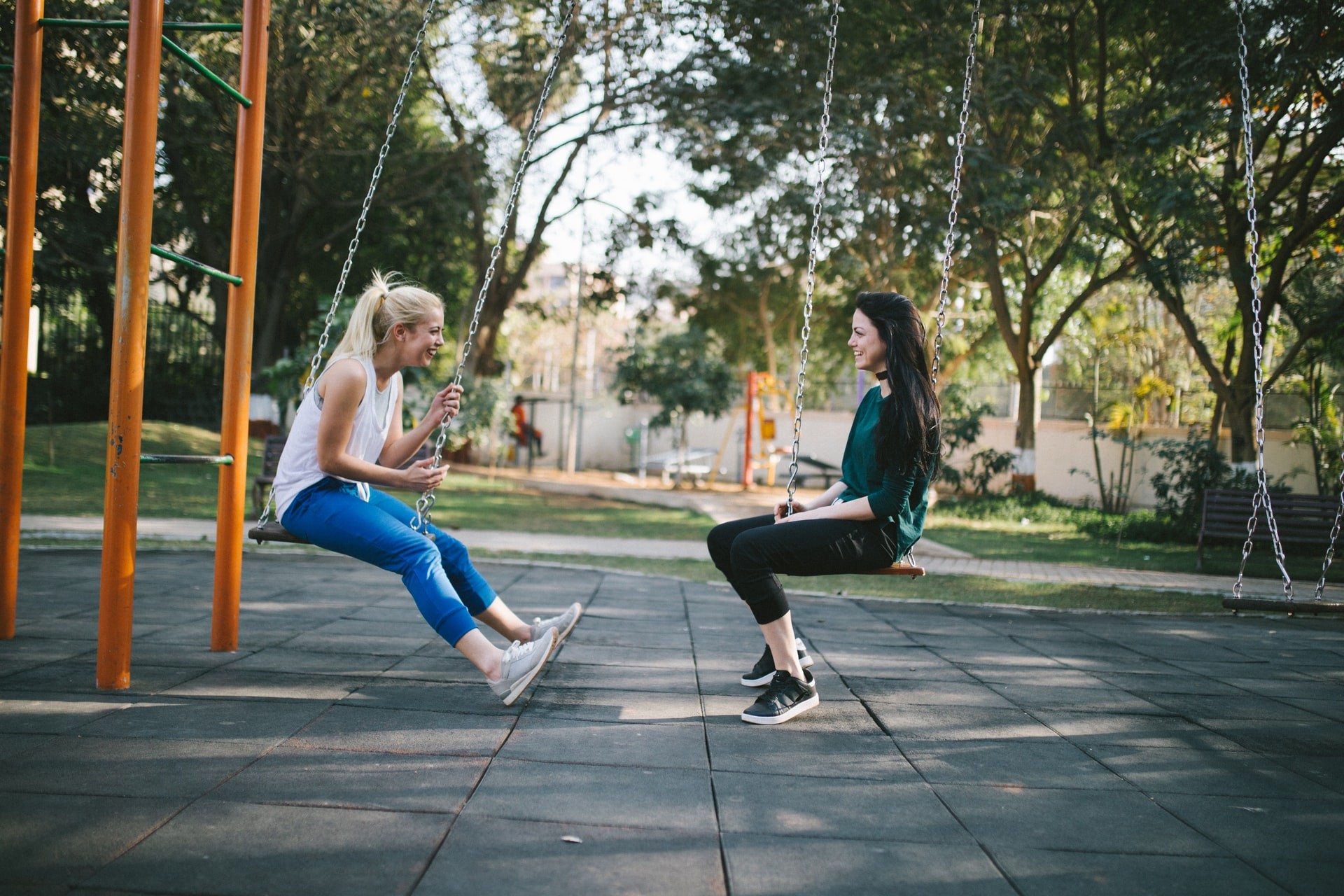 Two young women sit on swings in a playground, facing each other and smiling.