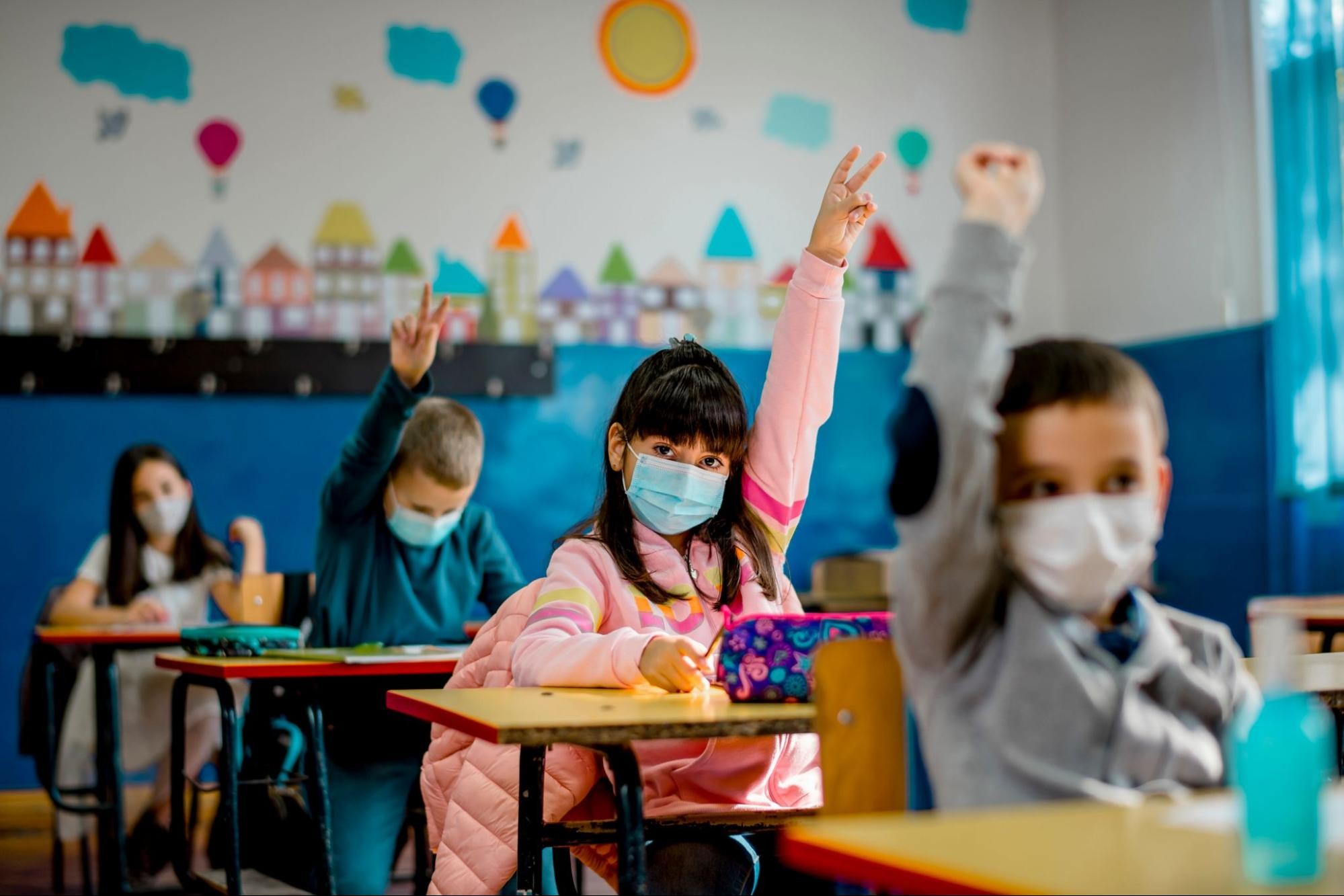 Four children sit in a row of desks, wearing masks and raising their hands, as they return to in-person learning after COVID-19.