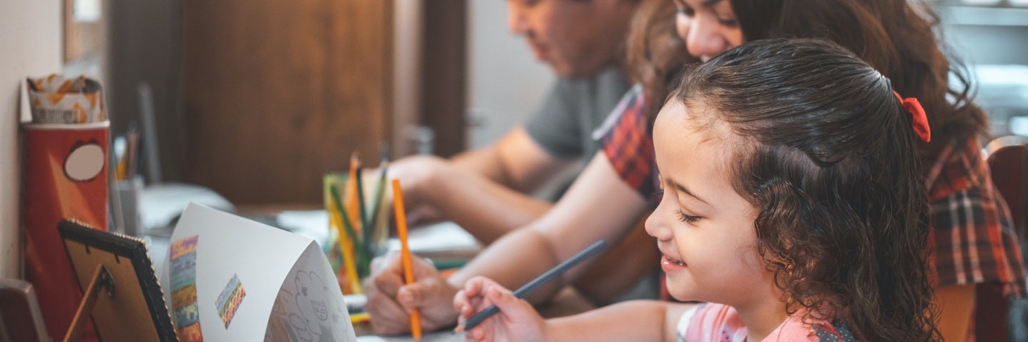 A father, mother and daughter sit at a table coloring something for spring break activities for kids.