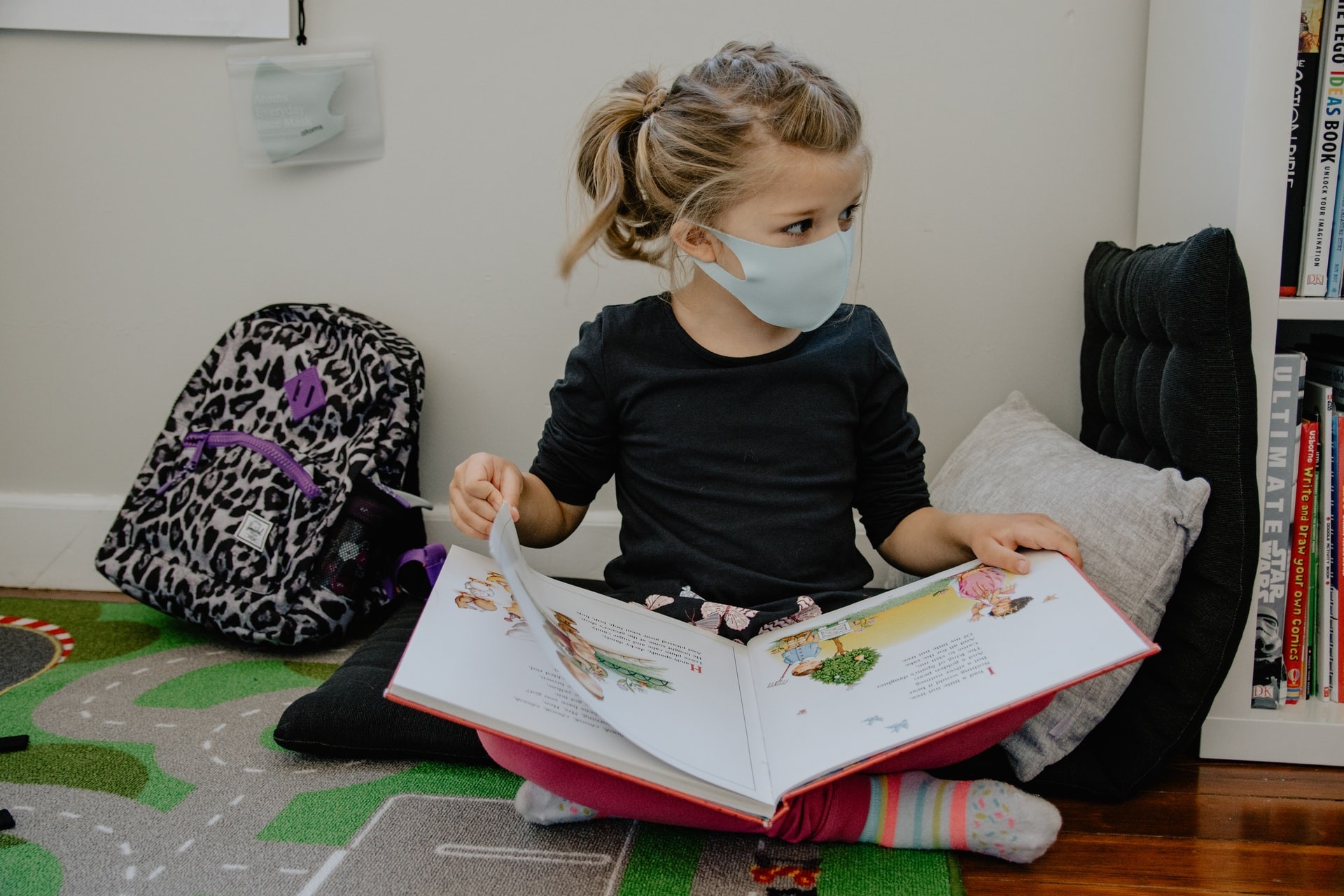 A young girl wearing a mask reads a book in a classroom. 
