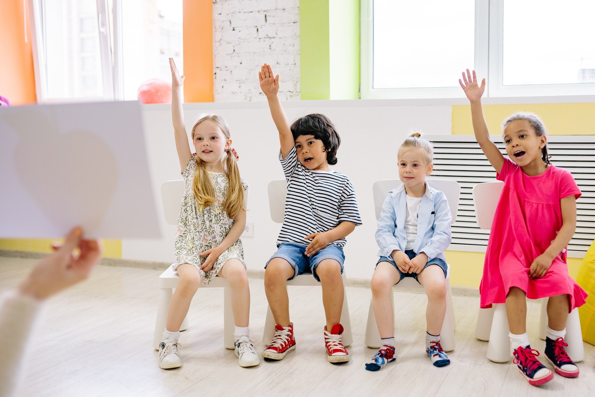 A row of students sit on a bench in the classroom and raise their hands.