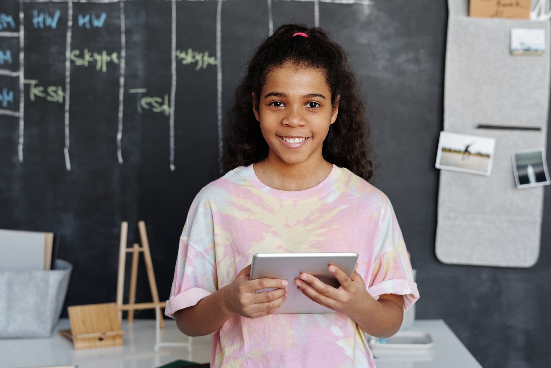 A young girl stands in front of a blackboard holding a tablet.