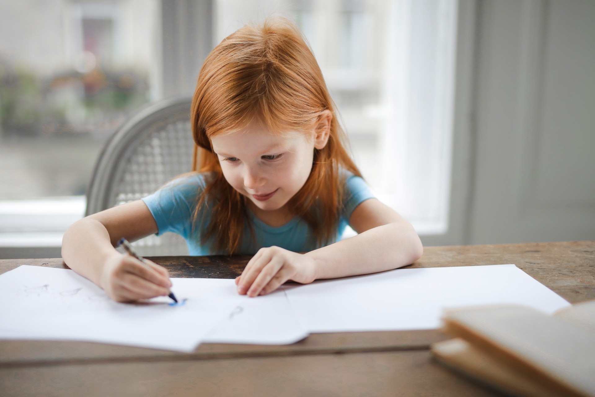 Young child sits at a table in front of a window while writing on a sheet of paper. 