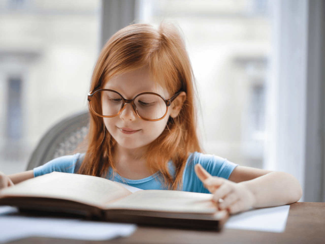 Child wearing glasses and smiling while reading a book at her desk.