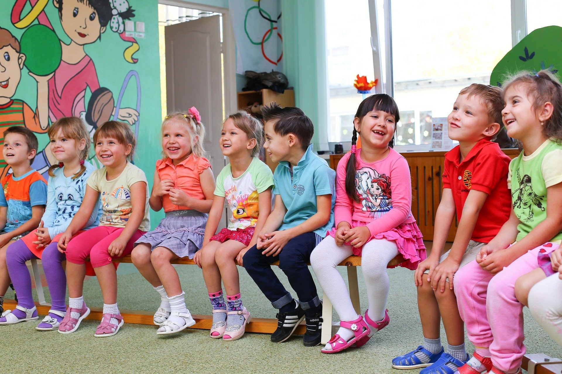 Nine kindergarten students sit in a row on wooden benches in a classroom.