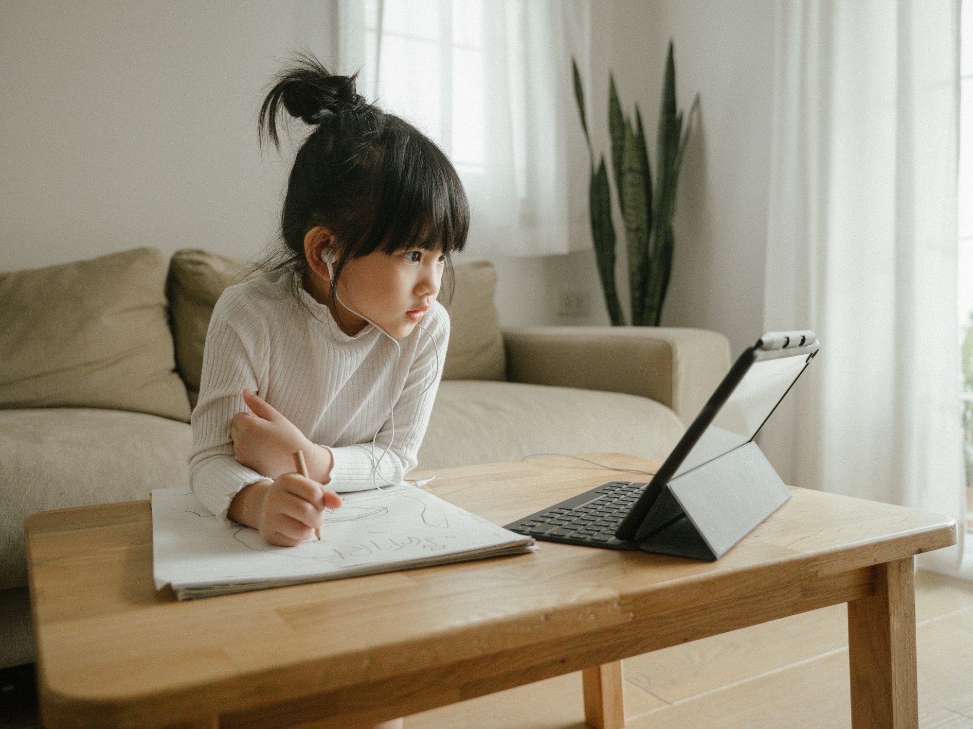 Young girl sitting at a table, watching something on her tablet while writing on a piece of paper with headphones in.