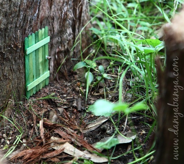Small green fairy door placed at the bottom of a tree trunk in a garden.