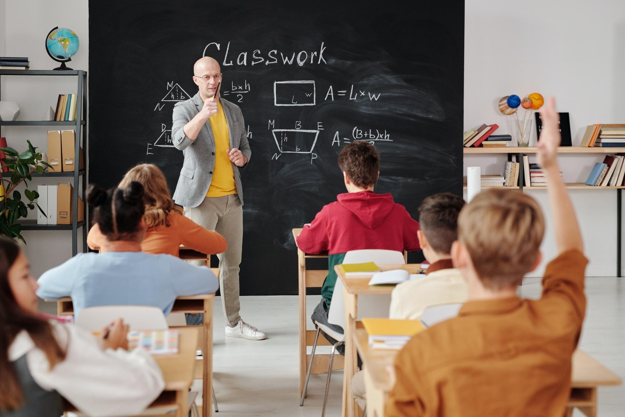 Teacher standing in a classroom in front of a blackboard, pointing at a student who is raising their hand.