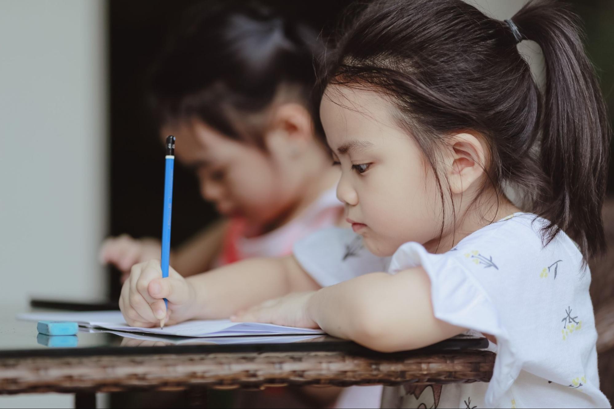 Two young girls write with paper and a pencil as they complete phonemic awareness activities in the classroom.