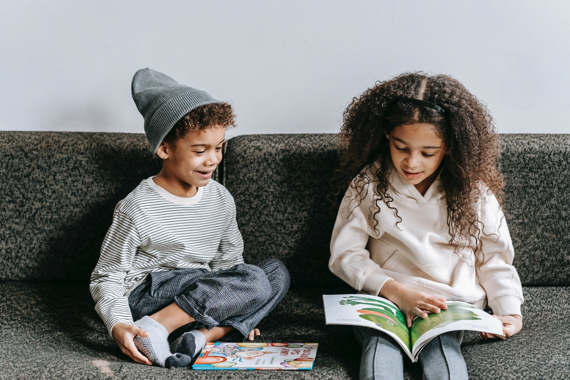 Two elementary aged children sit on a grey couch and read picture books for 1st graders.