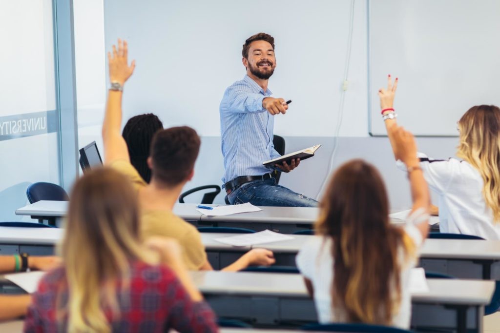 Teacher in front of a classroom and pointing at a student with a raised hand.