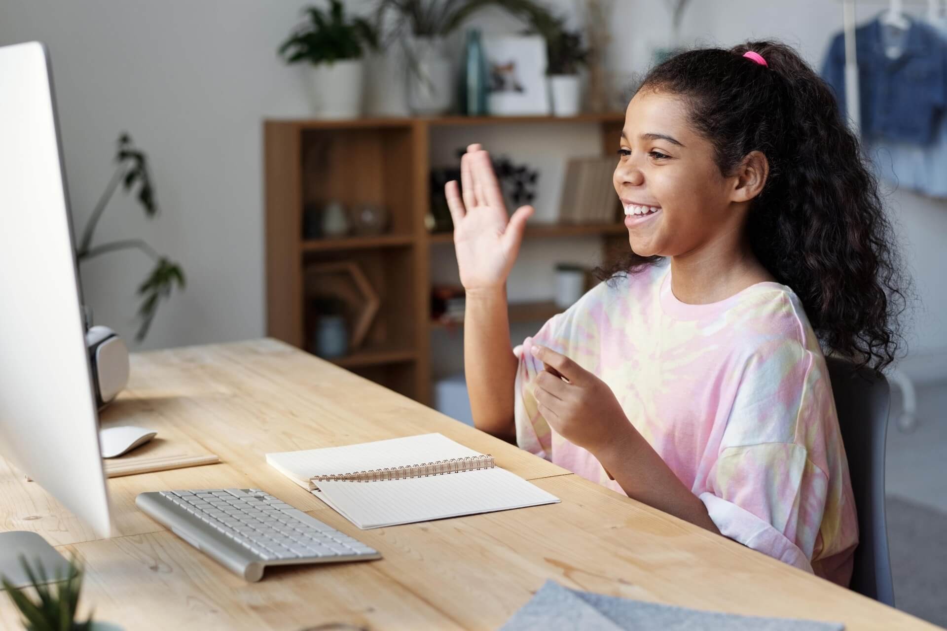 Young girl waves at a computer screen while participating in online learning. 