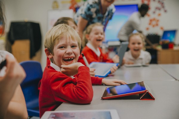 Young students sit smiling at a desk, using tablets to complete work.
