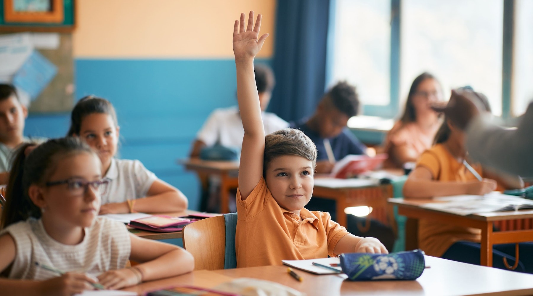 Child in classroom raising hand to answer a question.