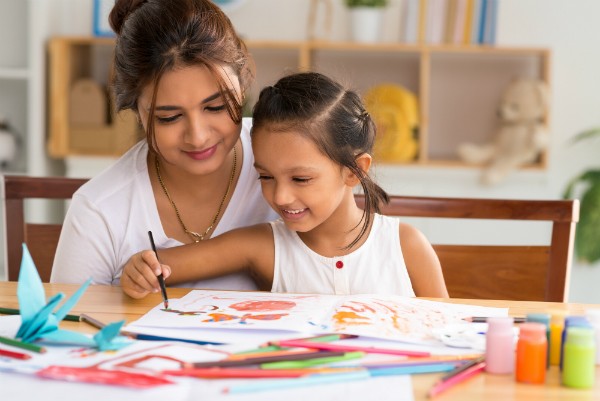 A mother and her daughter sit at a table at home coloring in a coloring book.