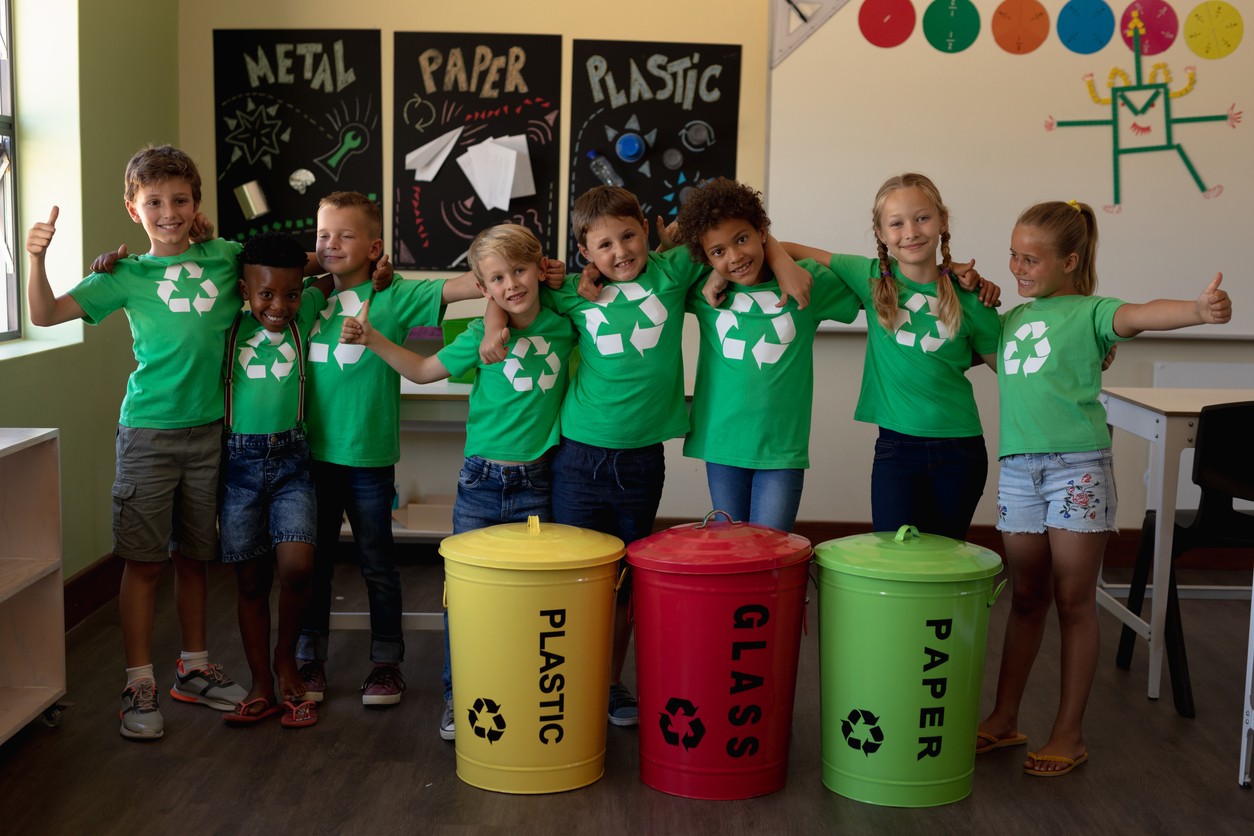 Eight elementary students stand in a row with arms around each other's shoulders wearing recycling symbol shirts and giving thumbs up behind plastic, glass and paper recycling bins.