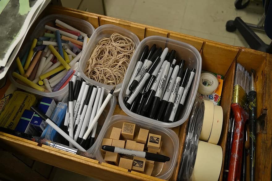 A teacher's organized desk drawer containing chalk, permanent markers, pens, stamps, tape and paint brushes.