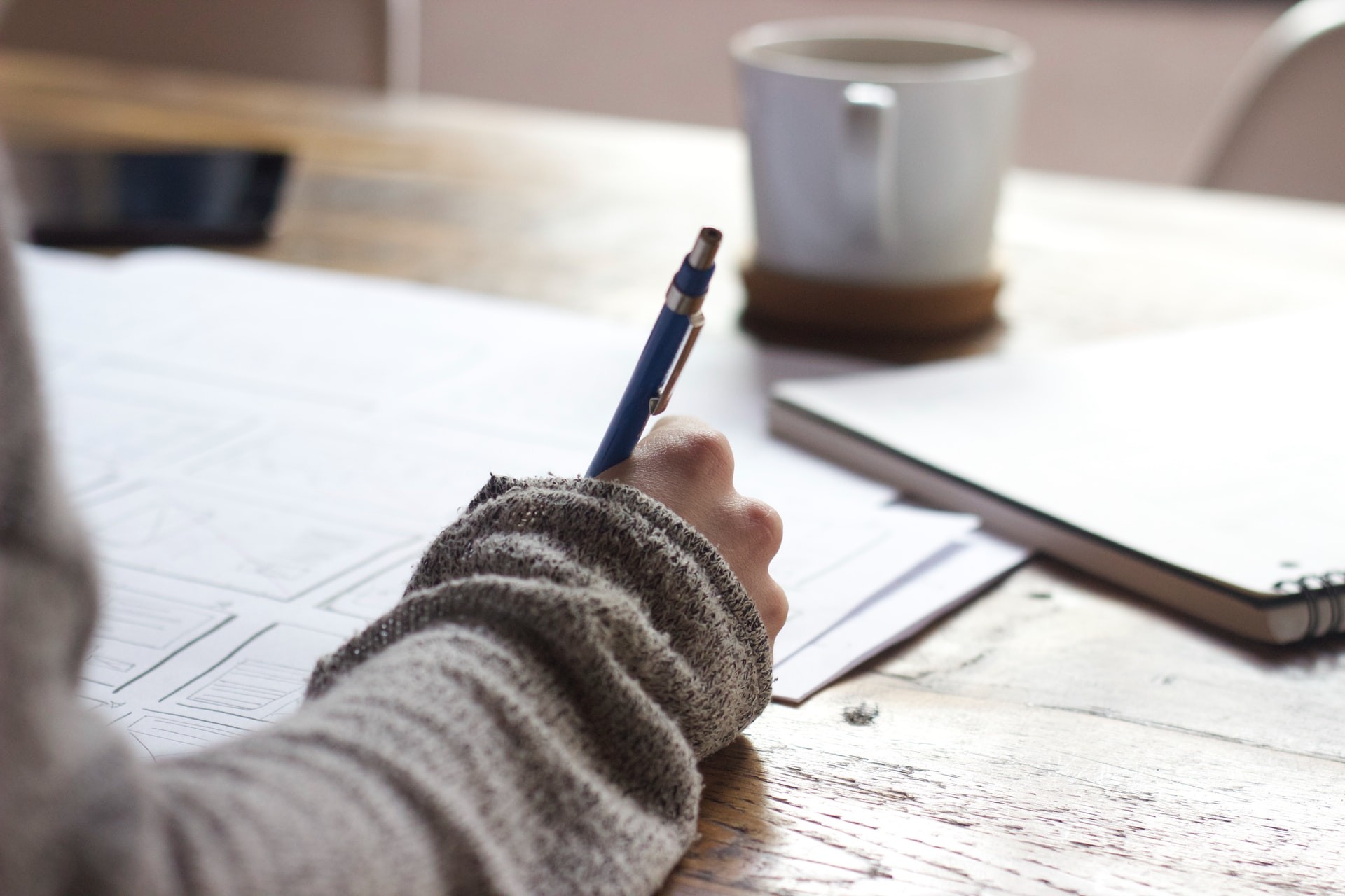 Teacher's hand writing a lesson plan with a notebook and a cup of coffee in the background.