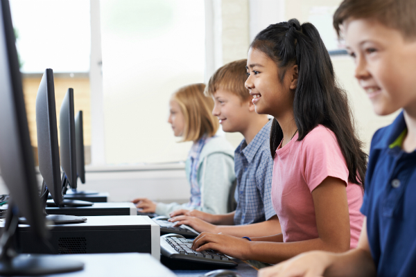 Four students sit in a school computer lab and work on desktop computers while smiling.