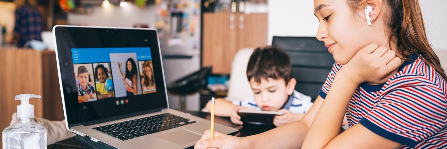 An older sister takes notes while in a classroom video call and her younger brother uses a mobile device.