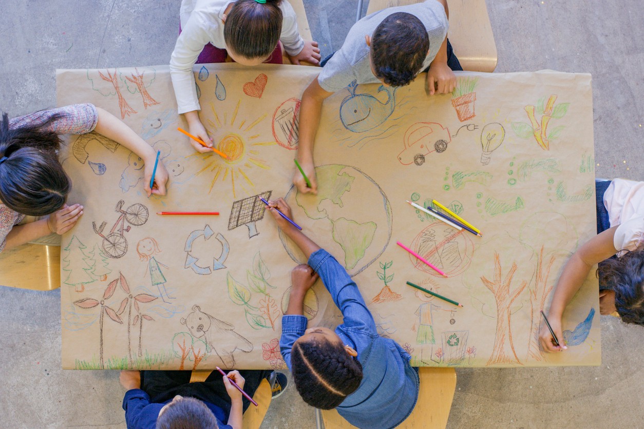 An aerial view of six students coloring Earth Day themed images on a table covered in brown paper.