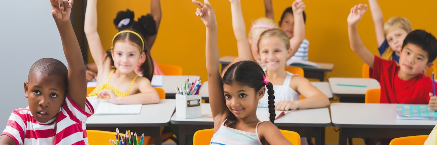 A class of young students sits at their desks and raises their hands