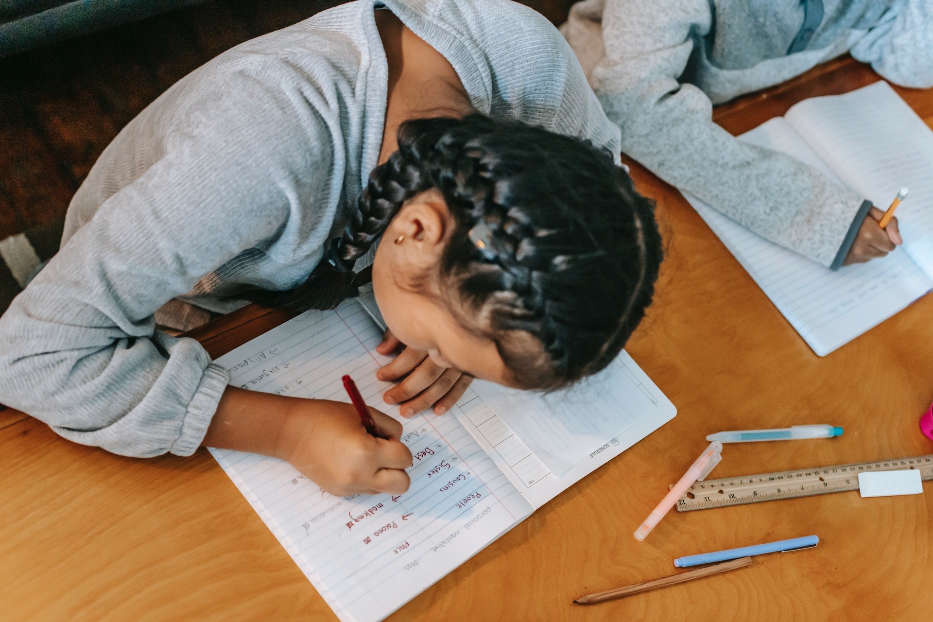 Top-down photograph of a girl with braids sitting at a desk next to another student and writing in a notebook.