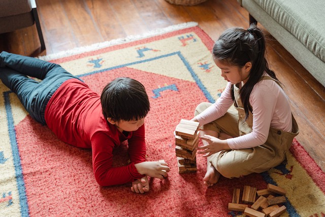 Two siblings laying on a carpet playing Jenga together.