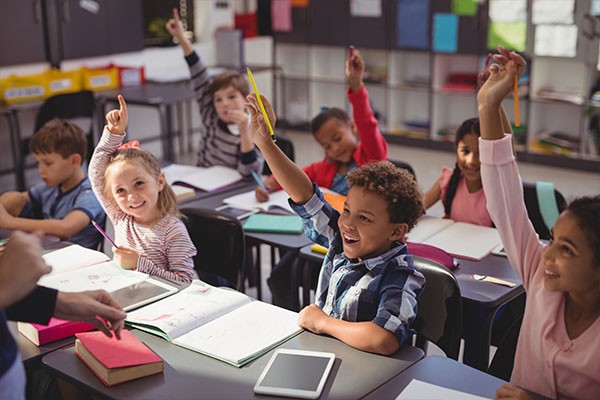 Young students sit in rows, raising their hands to answer the teacher's question.