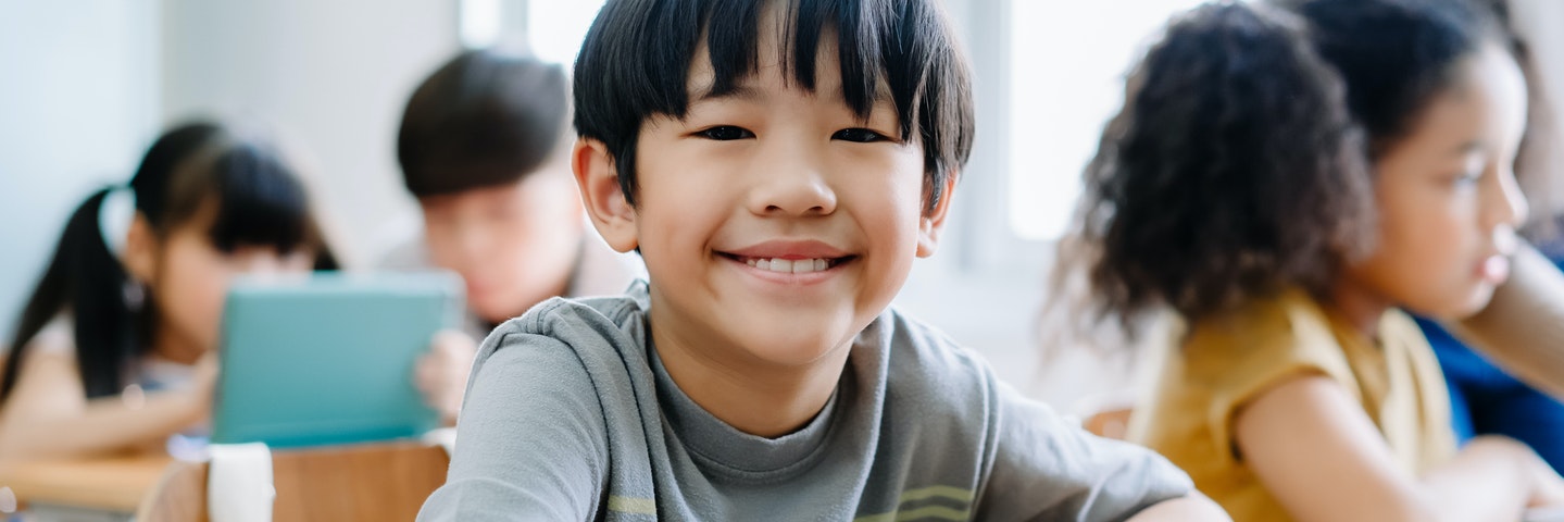 Young boy smiles at the camera while sitting at a desk with a tablet in a classroom.