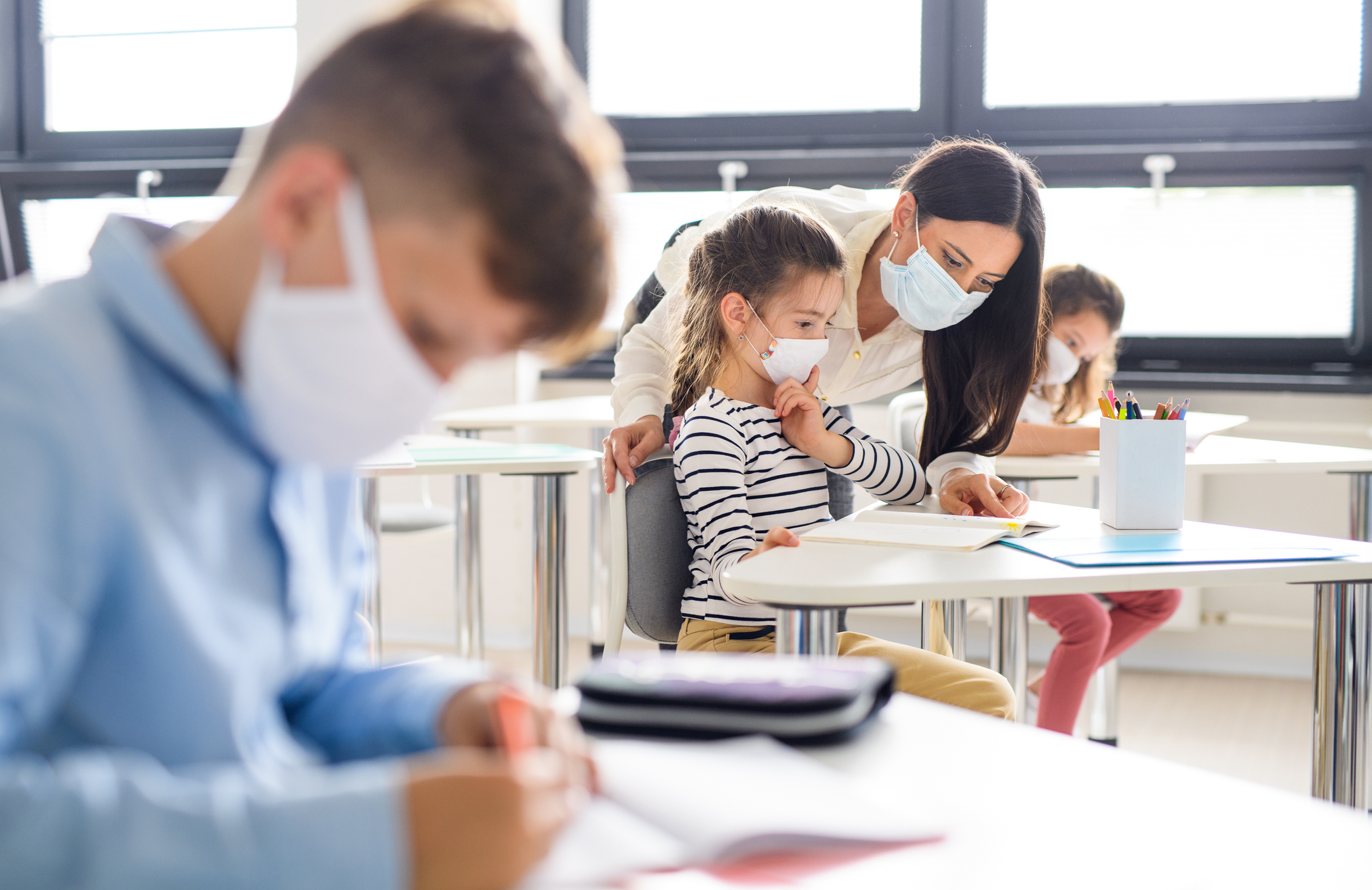 Three students sit socially distanced, wearing masks, in a classroom while a teacher helps one girl with her work.
